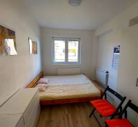 Garsonka s postelí, bílou skříní a podlahou s dřevěným dekorem, dvě sklápěcí židle.