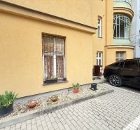 Parkování před budovou na Leškově v Bratislavě s květináči a zaparkovaným autem.