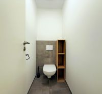 Toaleta s šedou dlažbou a dřevěnou policí ve 2-pokojovém bytě.