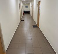 Úzká chodba v kancelářích s dlažbou, dveřmi a pohovkou na konci.