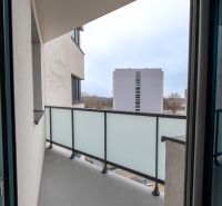 Balkon v 2-pokojovém bytě s výhledem na okolní budovy přes skleněné zábradlí.