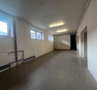 Dlouhá místnost s bílými stěnami, okny a litinovými radiátory ve skladech a halách.