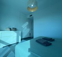 Rekreační apartmán se světlým pokojem, postelí, komodou a lodžií v modrých tónech.
