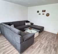 Obývací pokoj s rohovou sedačkou, skleněným stolkem a podlahou s dřevěným dekorem.