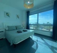 Ložnice rekreačního apartmánu s velkým oknem, postelí a stolky.