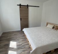 Ložnice v rodinném domě s postelí z palet, posuvnými dveřmi a podlahou s dřevěným dekorem.