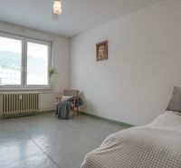 Ložnice v 2-pokojovém bytě s velkým oknem, křeslem a jednoduchým zařízením.