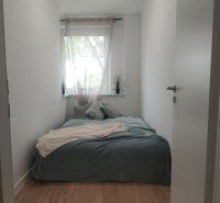 Úzká ložnice s postelí a oknem, podlaha s dřevěným dekorem, světlý interiér.
