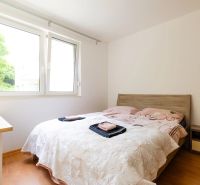 Ložnice v dvoupokojovém bytě s oknem, postelí a podlahou s dřevěným dekorem.