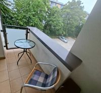 Balkon garsonky s výhledem na zeleň a parkoviště, místnost se stolem a židlí.
