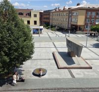 Náměstí SNP ve Zvolenu s monumentem a chodci, obklopené kancelářemi a stromy.