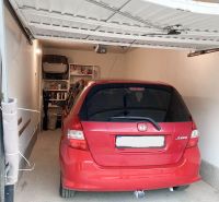 Červené auto zaparkované v garáži s regály na stěně, podlaha z dlažby.