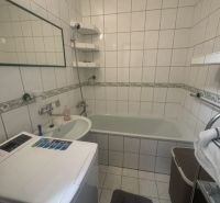 Koupelna v 2-pokojovém bytě s vanou, umyvadlem a pračkou, obložená bílými dlaždicemi.