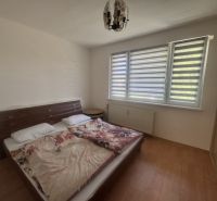 Ložnice v dvoupokojovém bytě s postelí, vedle okna se žaluziemi a skříní.