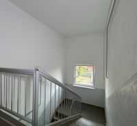Schodiště s oknem v 2-pokojovém bytě, stěny jsou bílé s kovovým zábradlím.