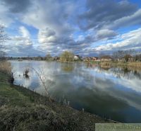 Poklidná vodní hladina s rodinnými domy v Tvrdošovcích a dramatická obloha.