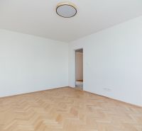 Prázdná místnost s dřevěným dekorem podlahy a výhledem do kuchyně ve 3-pokojovém bytě.