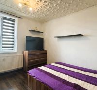 Ložnice ve 4pokojovém bytě s postelí, televizí, podlahou s dřevěným dekorem a moderními žaluziemi.