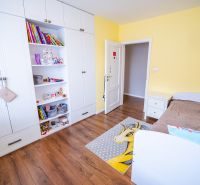 Dětský pokoj v rodinném domě s bílou skříní, hnědou postelí a podlahou s dřevěným dekorem.