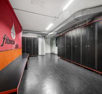 Fitness šatna s černými skříňkami a červenou stěnou v obchodních prostorách.