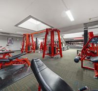 Fitness centrum s posilovacími stroji a červenými lavičkami, vhodné obchodní prostory.
