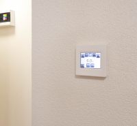 Nastavitelný termostat na stěně v garsonce, jednoduchý design, bílé pozadí.