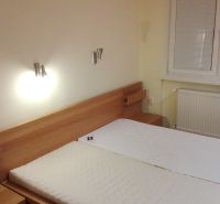 Ložnice třípokojového bytu s dřevěnou postelí a oknem s roletami na stěně.