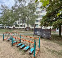 Exteriér bytovky v Topoľčanech s dětským hřištěm a vegetací kolem 1-pokojového bytu.
