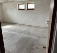 Nevybavená místnost v rodinném domě se dvěma malými okny a surovou podlahou.