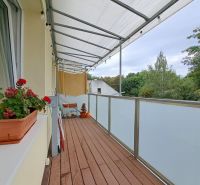 Balkon v 3-pokojovém bytě s podlahou s dřevěným dekorem, květinami a výhledem.