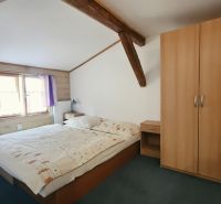 Ložnice v rekreačním apartmánu s dřevěnými trámy, skříní a postelí vedle okna.