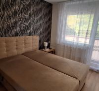 Ložnice s čalouněnou postelí, vzorovaná tapeta, záclony, podlaha s dřevěným dekorem.