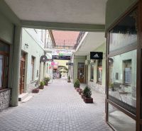 Pasáž s obchodními prostory v Topoľčanech se stromečky a cedulkami na budovách.