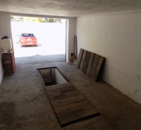 Garáž s montážní jámou, starší betonový povrch a zaparkované auto venku.