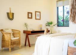 Světlá ložnice v rodinném domě s ratanovým křeslem a dřevěným stolem.