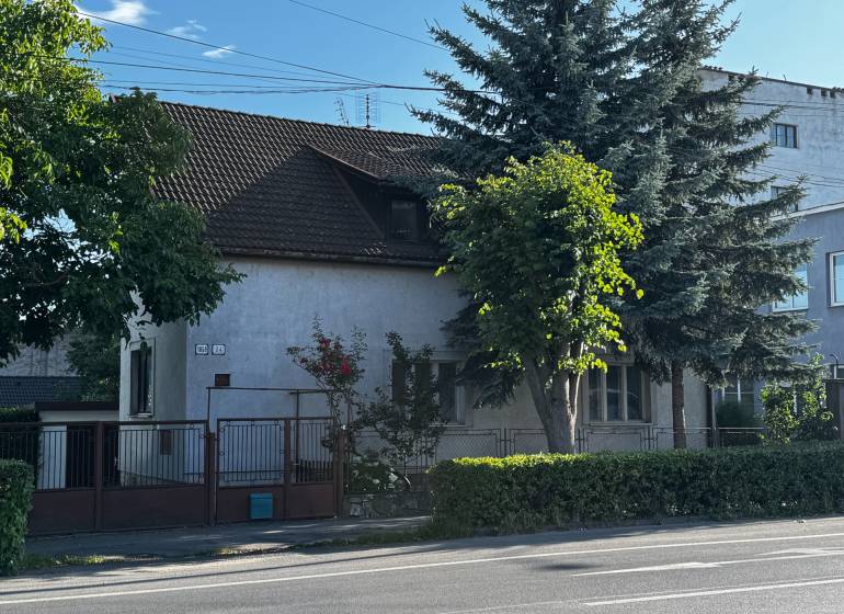 Rodinný dům v Lučenci obklopený stromy a živým plotem, s přístupem z hlavní ulice.