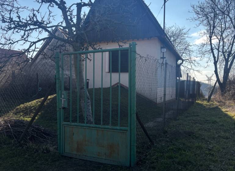 Malá chata obklopená stromy, oplocená zahrada, ideální pro rekreační pozemky.