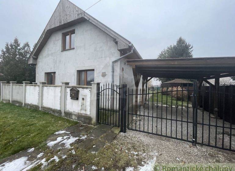 Rodinný dům s bílou fasádou a bránou, obklopený trávníkem a oplocením.