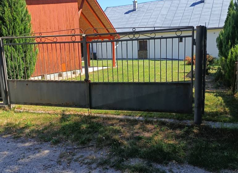 Brána a dřevěná chata v Tužině, obklopené trávníkem a jehličnany.