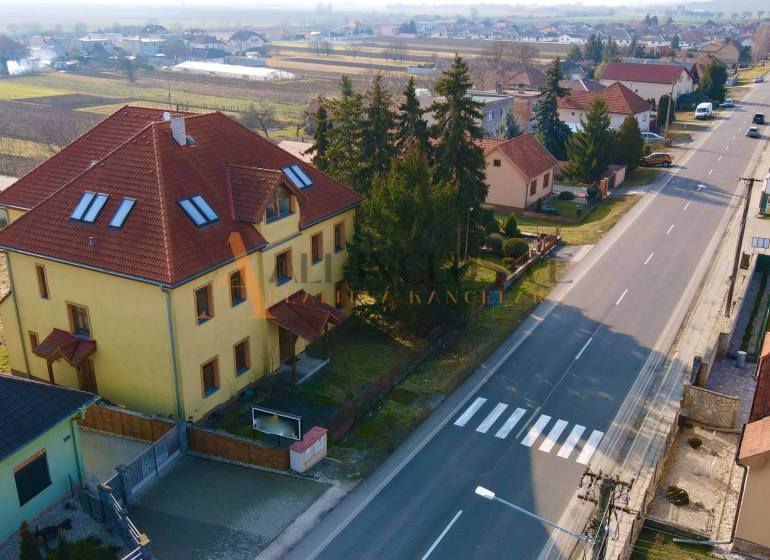 Budova na ulici Chrabrany v Topoľčanoch, obklopená domy a stromy, u hlavní silnice.