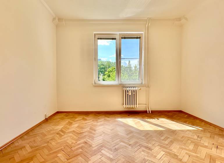 Prázdný pokoj v třípokojovém bytě s parketami a oknem s výhledem na zeleň.