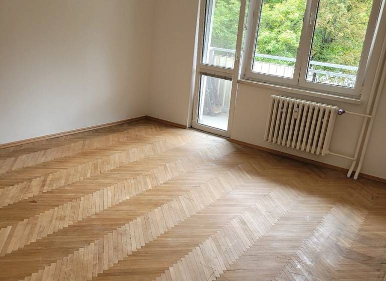 Pokoj ve 3-pokojovém bytě s dřevěnou podlahou a balkonovými dveřmi.