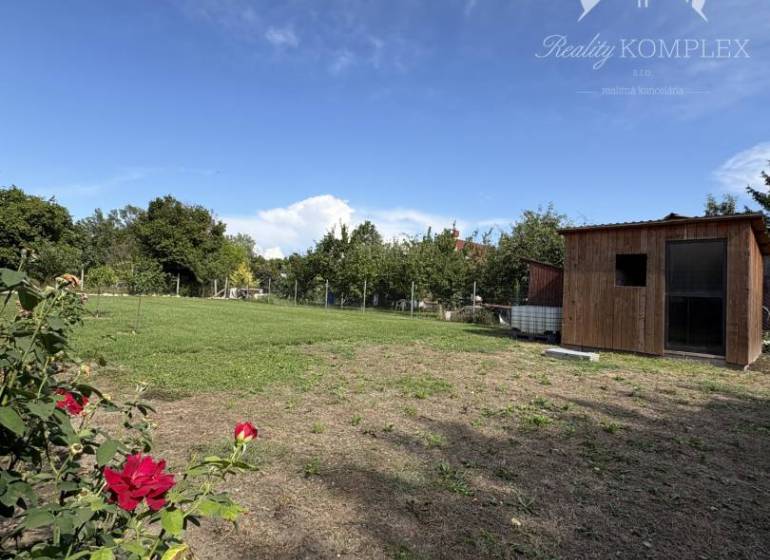Zahrada u chaty v Komárně s dřevěnou budovou a červenými květy.