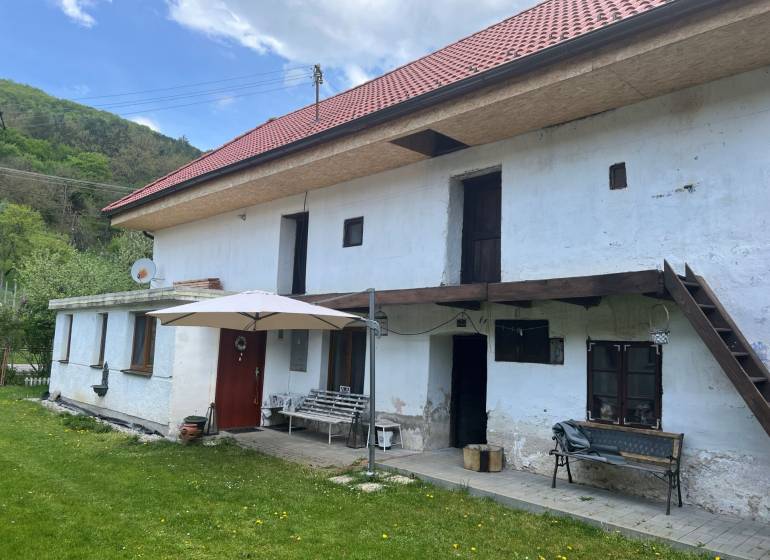 Venkovská chata s červenou střechou a terasou, obklopená zeleným trávníkem a přírodou.