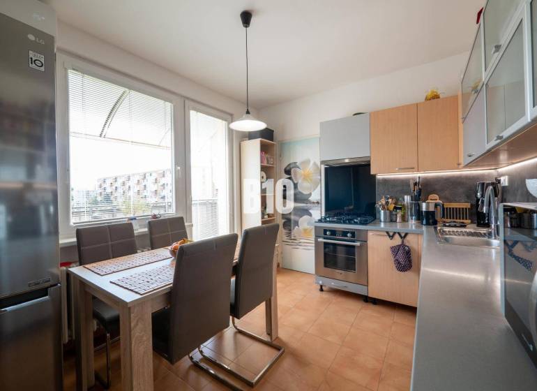 Kuchyně ve 4-pokojovém bytě s jídelním stolem, hnědými židlemi a velkým oknem.