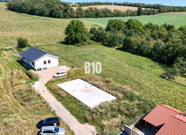 Záběr na pozemek pro bydlení v Čeľadicích zahrnuje dům, zeleň a auta.