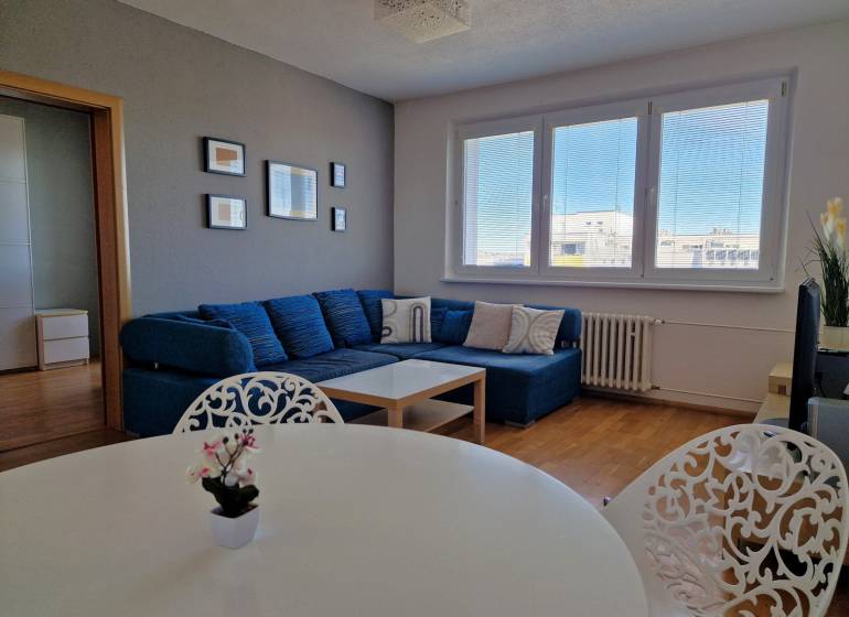 Obývací pokoj 3-pokojového bytu s pohovkou, bílým stolem a podlahou s dřevěným dekorem.