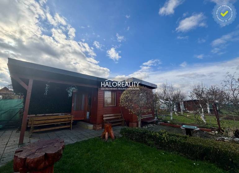 Dřevěná chatka v zahradách Spišské Nové Vsi s lavičkami a květinovým záhonem.