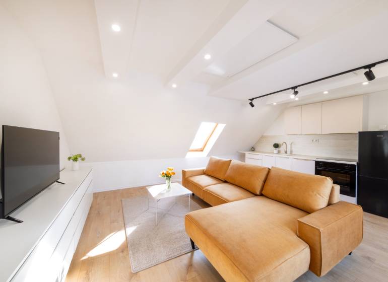 Obývací pokoj s kuchyní ve 2pokojovém bytě: velké okno, hnědá sedačka, podlaha s dřevěným dekorem.