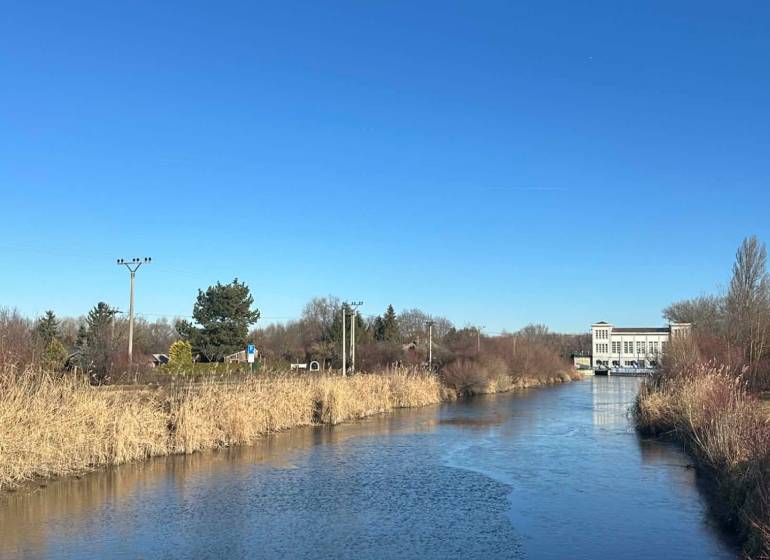 Řeka obklopená rákosím v Zahradách na Nové osadě v Komárně pod modrou oblohou.